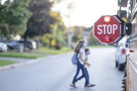 A flashing school bus stop sign in focus with students boarding in the background.